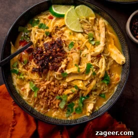 Overhead picture of a black textured bowl full of chicken laksa topped with some chilli crisp and a sprinkling of fresh coriander and some lime wedges.