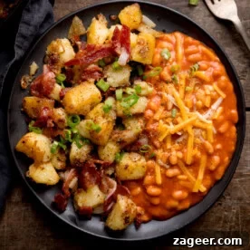 A black plate with baked beans topped with grated cheese on one side and a hearty potato and bacon hash with potatoes, bacon pieces, and chopped spring onions on the other.